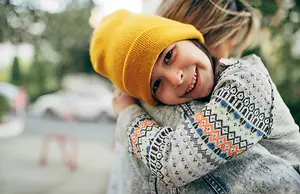 A little girl wearing a yellow beanie is being hugged by a woman, and both are smiling.