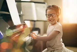 A young girl with glasses playing a piano with books and a music stand in the background