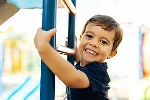 A smiling boy with braces is holding onto a blue bar on a playground equipment in an outdoor area.