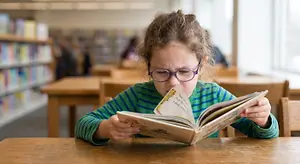 A young girl wearing glasses is reading a book in a library with a serious expression on her face