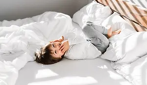 A child is lying on a bed with white sheets and pillows, smiling and covering his mouth with his hand. The bed has a wooden headboard and the child's shadow falls on the bed. The room is illuminated by sunlight.