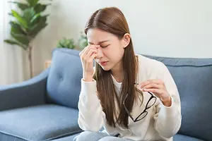 A woman sitting on a couch with her hand on her forehead and holding her glasses