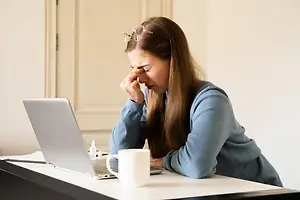 A woman with glasses on her head and a blue sweater covering her face with her hand and holding a white mug on the table in front of her.