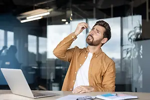 A man with a beard is sitting at a desk in an office, holding his nose, looking at something with his mouth open.