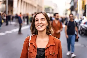 A woman with a smile on her face standing on the street, with people walking behind her.