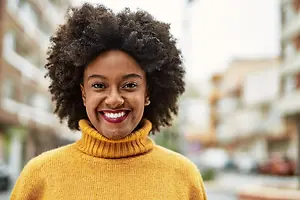 African American woman with curly hair wearing a yellow turtleneck sweater standing on a city street smiling at the camera