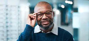 A smiling man with a beard wearing glasses and a blue sweater is standing in a room, holding his glasses with his right hand while looking at the camera and smiling.