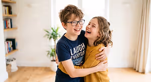 Two children, a boy wearing glasses and a girl, are hugging each other and smiling in a room with wooden floors and shelves with books, a plant, and a window with curtains.
