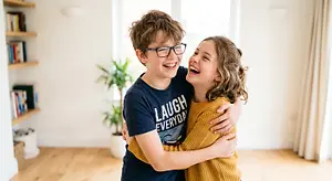 Two children, a boy wearing glasses and a girl, are hugging each other and smiling in a room with wooden floors and shelves with books, a plant, and a window with curtains.