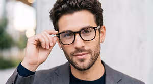 A man wearing a suit, glasses, and smiling while holding his glasses
