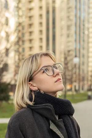 A woman with blonde hair wearing a black coat and glasses is standing and looking up at the sky in front of a blurry building.