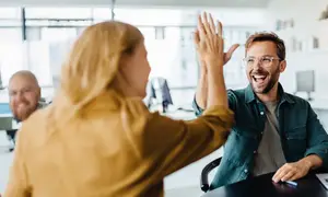 A woman and a man are giving a high-five gesture in a room with glass windows and shelves in the background