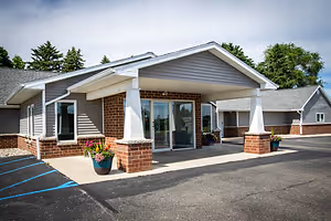 The entrance to a building with a gray roof, white trim, brick walls, glass doors, and windows, with a gray pavement, a blue line, and two potted plants in front.