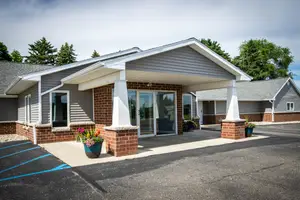 The entrance to a building with a gray roof, white trim, brick walls, glass doors, and windows, with a gray pavement, a blue line, and two potted plants in front.