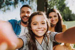 A young girl is taking a selfie with her parents in a park.
