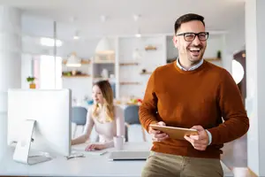 A man holding a tablet with a smiling face is standing in front of a desk with a woman sitting behind him, both looking at the monitor. Behind them is a cabinet with shelves and a potted plant on top.