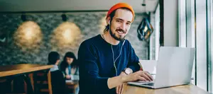 A man wearing an orange hat and earphones is working on his laptop while sitting at a table in a cafe.