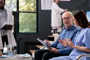 An older man speaks with a female doctor in a clinic office while a male doctor walks up the stairs behind them