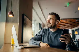 A man in a gray shirt sits at a table in a restaurant and holds a phone.