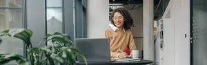 A woman sitting in an office with a laptop on a desk, smiling and looking at the camera.