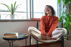 A woman sitting on a chair inside her home, smiling while looking out the window, next to a table with books and a phone, and a plant in a white pot behind her.