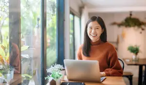 A woman is sitting at a desk with a laptop and smiling while looking at the camera