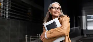 A woman wearing glasses and a brown coat stands with a folder in her hand