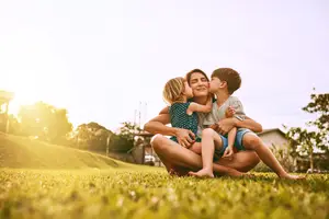 A mother is sitting on the grass with two children and they are kissing each other and smiling