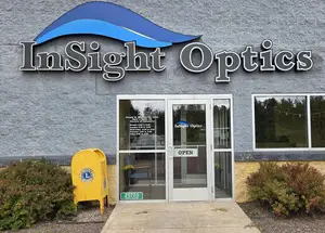 The front entrance of InSight Optics, a building with gray walls, glass doors, and a sign displaying the business name in blue and white letters, accompanied by a yellow mailbox and greenery on either side.