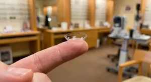 A finger is holding a contact lens in a room with blurry background.