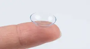 A fingertip holds a transparent contact lens with a reflection of light