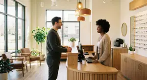 A man and a woman are standing in front of a counter, and the woman is smiling and seems to be explaining something to the man. The counter has a monitor, a keyboard, and a mouse. Behind them, there is a desk with a plant, a chair, and a couch. There are also lamps hanging from the ceiling and a shelf with eyeglasses on the right.