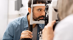 A man wearing a blue shirt is using a slit lamp to examine his eyes with an eye doctor behind him in a medical setting.
