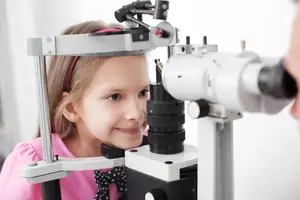 A young girl wearing a pink shirt is having her eyes checked by a doctor using a device.