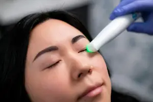 A woman with brown hair is undergoing a procedure where her eyes are closed, and she is being treated with a device emitting green light. A person in blue gloves holds the device, which is pointed at the woman's face.