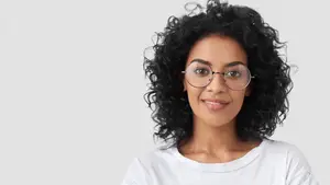 A smiling woman with curly hair wearing glasses and a white t-shirt standing against a white background