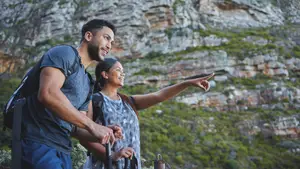 A man and a woman are standing near a cliff with green grass and rocks and they are pointing at the cliff