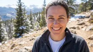 Woman wearing a fleece jacket and white shirt standing on a rocky trail in front of trees and mountains