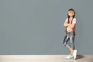 A young girl with glasses and a pink shirt stands confidently with her arms crossed, wearing silver patterned pants and white sneakers, in a room with a gray wall and a wooden floor.