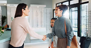 Two adult women with a young girl smiling and shaking hands in front of a counter with a woman wearing glasses and a handbag behind them
