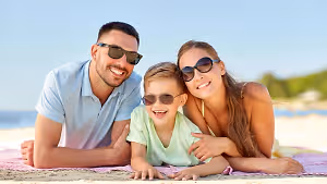 A man, a woman, and a boy are lying on a beach with sunglasses on, smiling and posing for a photo