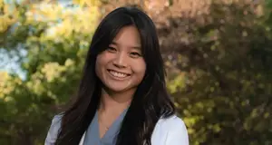 Portrait of a woman with long black hair wearing a white coat and smiling for a photo against a background of blurred trees