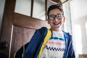 A boy with braces smiles and stands in front of a wooden door, wearing a backpack and glasses.
