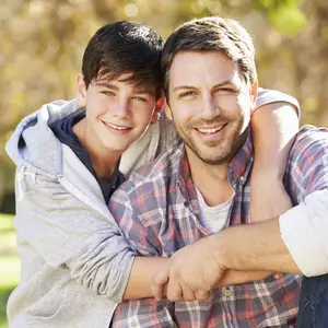 A smiling father and son pose for a photo, with the father wrapping his arms around the boy's shoulders, both looking at the camera.
