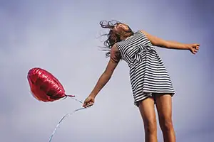 A woman in a black and white striped dress is flying a red heart-shaped balloon in the sky