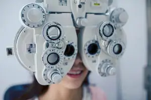 A woman in a pink shirt looks through a white phoropter during an eye exam.
