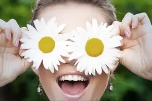 A woman is holding two daisies in front of her face, smiling, and showing her teeth with a blurry background behind her.