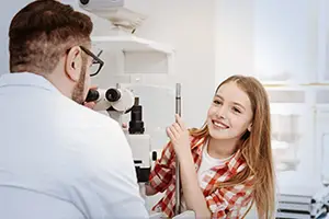 A young girl is smiling and looking into an ophthalmoscope while an adult male is examining her in a clinic