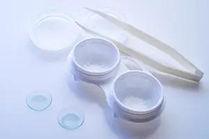 Close up of contact lenses, lens case, and tweezers on a white surface