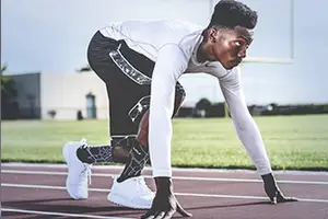 A man wearing a white long-sleeve shirt and black shorts is standing on a track, with his hands on the ground and his legs slightly apart, ready to run. He is wearing a black and white sock with a black and white design on it, and he is wearing white sneakers with black laces.